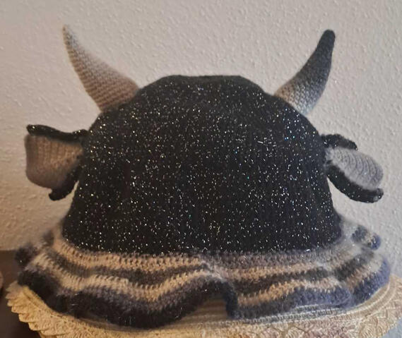 Dragon bucket hat A crocheted bucket head in glittering black, blueish-grey and beige. It has small horns at the top and floppy fin-like ears on the sides.